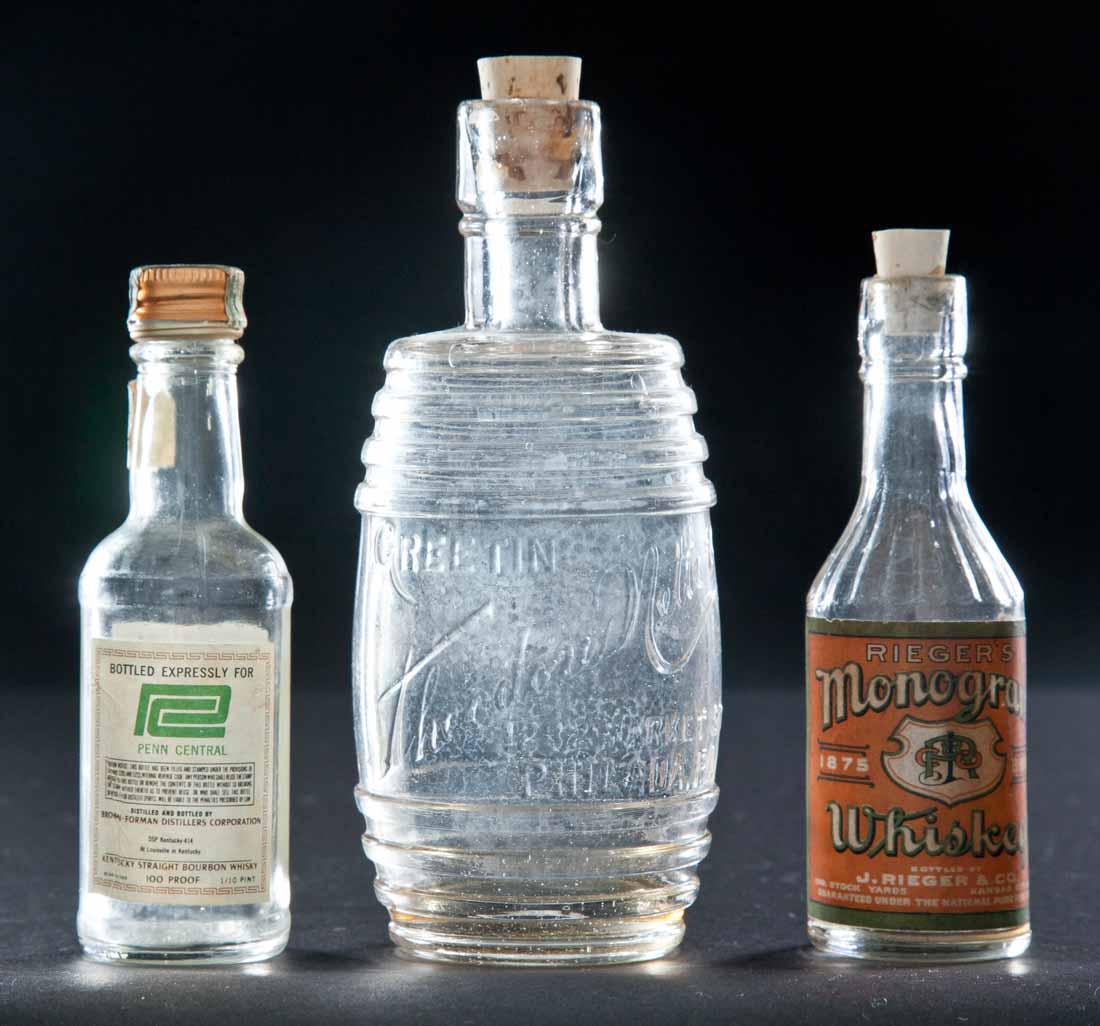 THREE COLORLESS MINIATURE BOTTLES: Barrel-shaped bottle with embossed label, "Greetings from Theodore Netter, 1232 Market St., Philada. PA.". 5.75"h. Airplane bottle with original label, "Rieger's Monogram Whiskey". 4.5"h. Airplane-siz