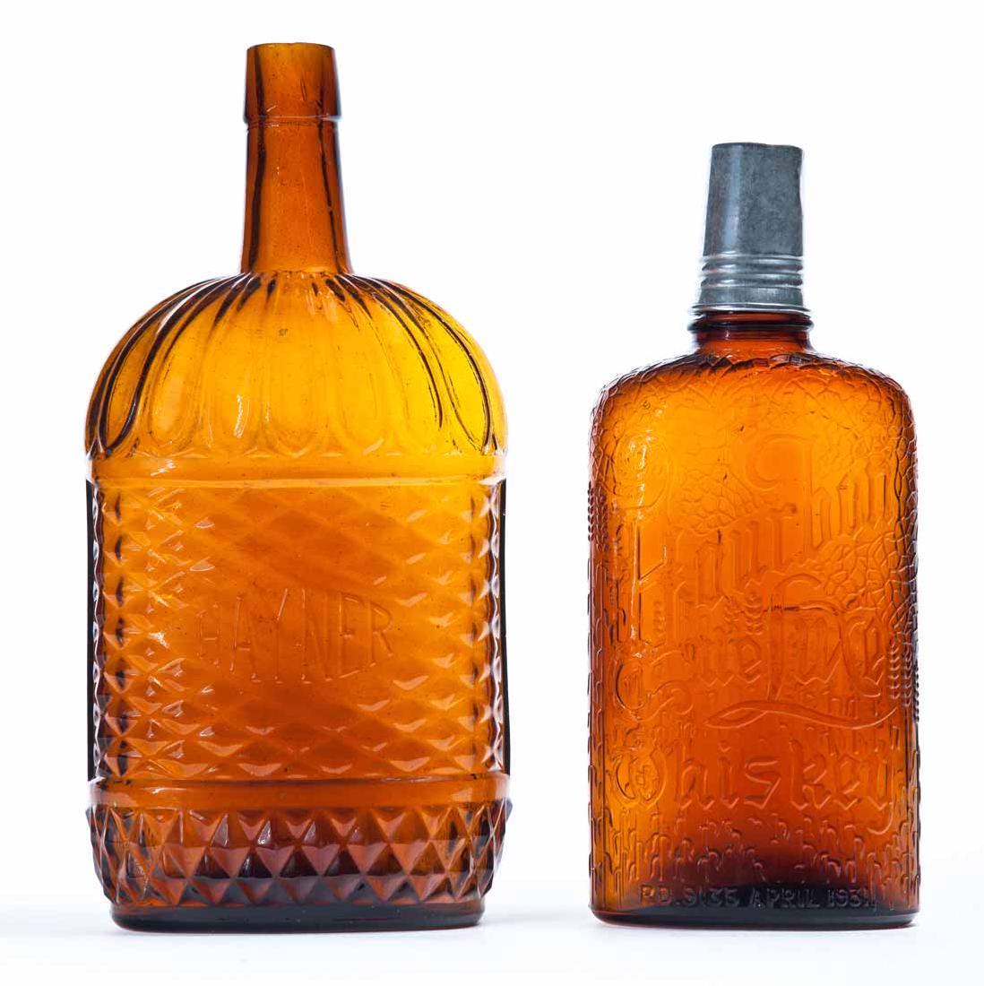 TWO AMBER WHISKEY FLASKS: Both pre-Prohibition with embossed labels. One for "United Distillers Bourbon deLuxe Whiskey, April 1931" with original cap and shot glass. 8.75"h. One "Hayner". 10"h.