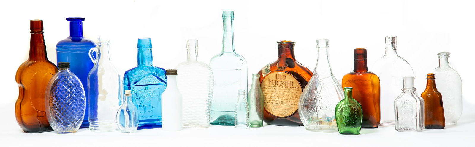 GROUP OF GLASS BOTTLES: American, late 19th-early 20th century. Group of nineteen glass bottles and flasks, two cobalt, three dark amber, one emerald green with "Benjamin Franklin", two aqua, one milk glass and nine clear. 3