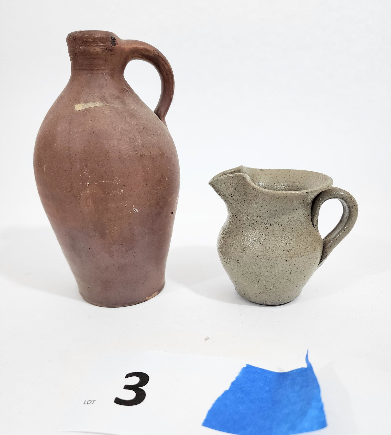 TWO MINIATURE PIECES OF POTTERY: American, mid-19th century. Includes an ovoid earthenware jug with a rust-color gaze, 7.25" high; together with a stoneware pitcher with an exaggerated rim and spout, 3.5" high.