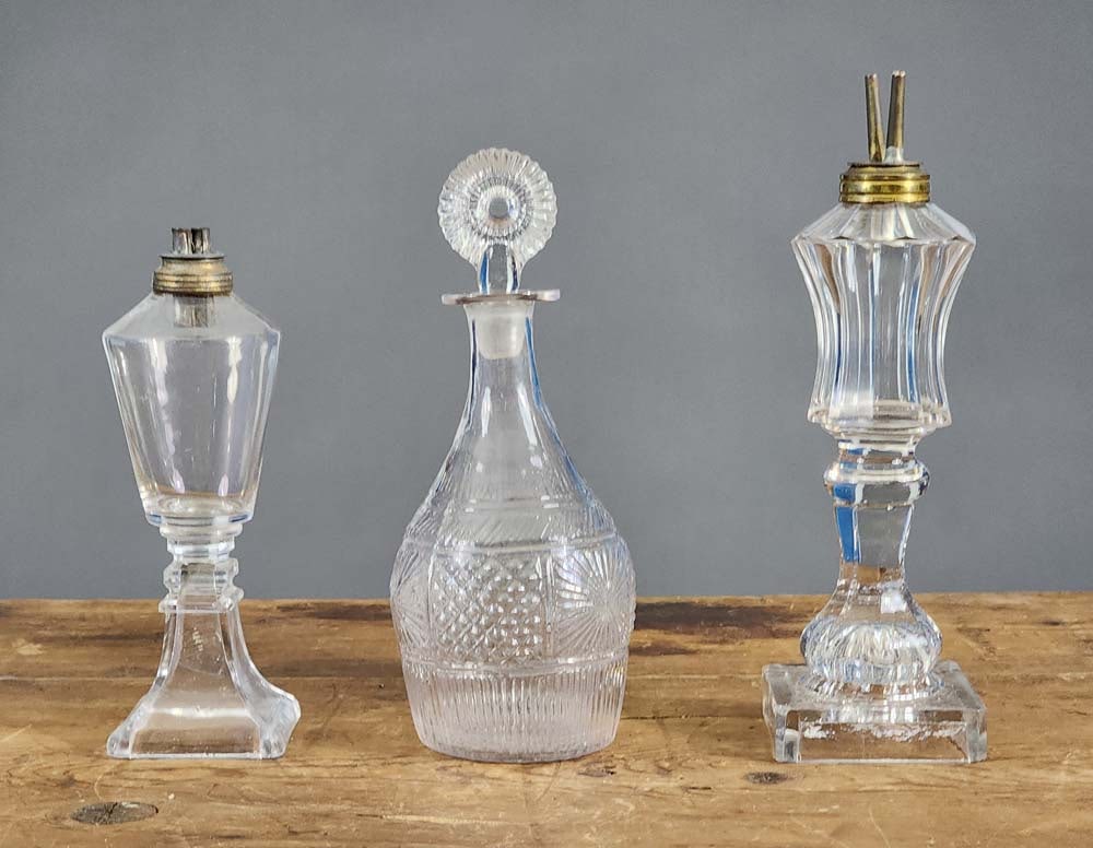 TWO EARLY FLUID LAMPS AND A DECANTER: Mid-19th century, colorless glass. Includes a flint glass camphene lamp, 10.5" high; together with a smaller whale oil lamp, 8.25" high, and a Boston and Sandwich mold-blown decanter, 9" high.
