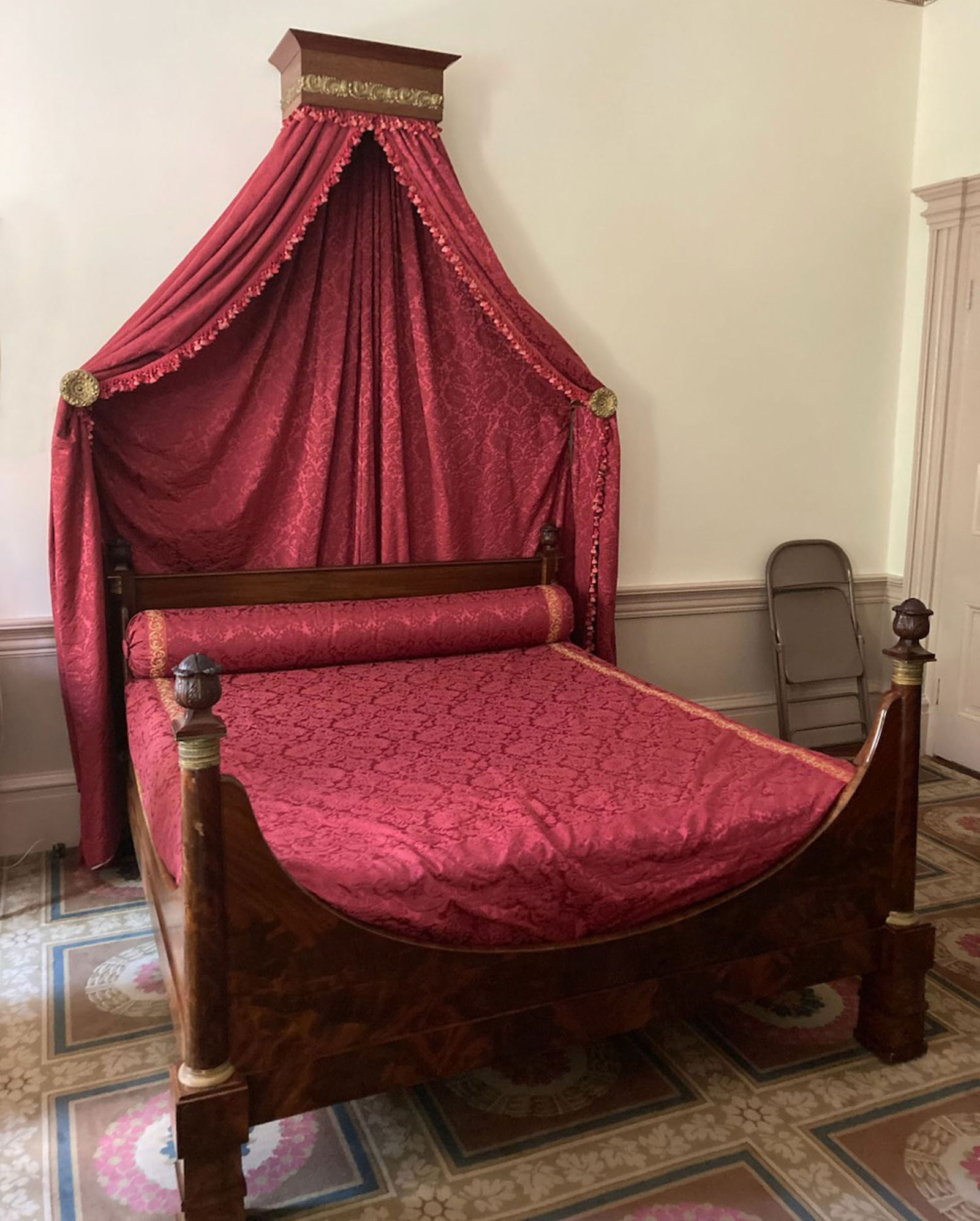 REGENCY BEDSTEAD: Probably England, c. 1830, mahogany. Figured veneer on the headboard, footboard, and rails, with blocked feet, carved finials, and ormolu mounts. 42.75" high, 66" wide, 78.5" long. With wall-mounted c