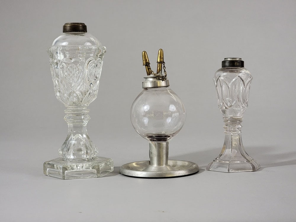 THREE PIECES OF EARLY GLASS LIGHTING: American, early-mid-19th century. Includes two colorless pressed oil lamps, and a colorless peg lamp in a pewter chamberstick base by Smith and Company, Boston. Tallest is 8.25" high. Provenance: a Mi