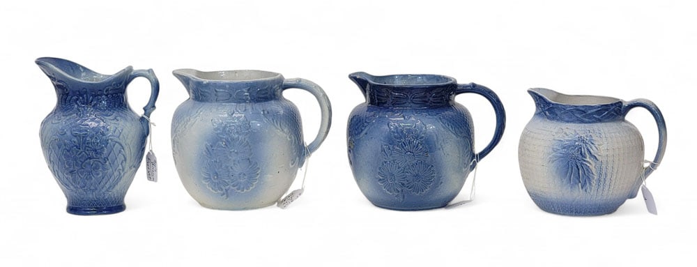 FOUR MOLDED PITCHERS WITH FLOWERS: American, early 20th century, stoneware. Four molded pitchers with flower designs, unmarked. One short round pitcher with "P"ointsettia, blue on white ground. 6.5" high; Two short round pitchers with