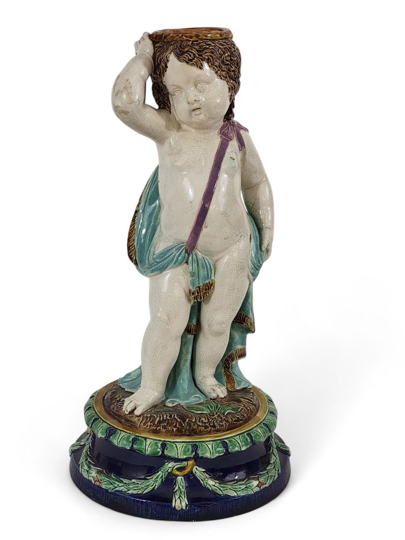 MAJOLICA PUTTO LAMP BASE: Probably Holdcroft, English, late 19th century, earthenware. Majolica lamp base depicting a bored-looking putto with turquoise sash or cape, over a pedestal with cobalt ground and green foliate garlan