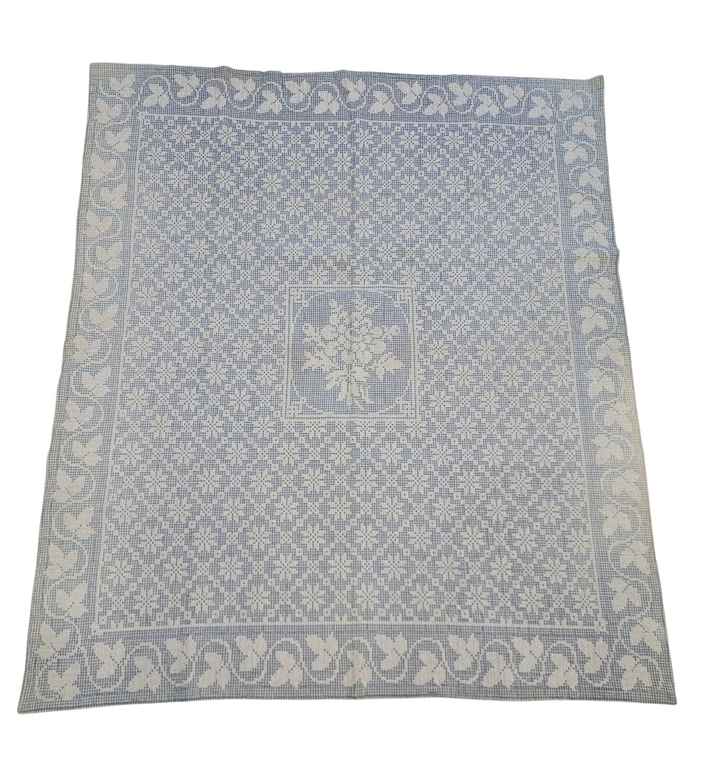 CROSS-STITCH QUILT: American, early 20th century, cotton. An embroidered quilt composed of white cross-stitch tiles on a blue gingham ground, with cross-stitch ivy vine border and a central cross-stitch flower arrangemen