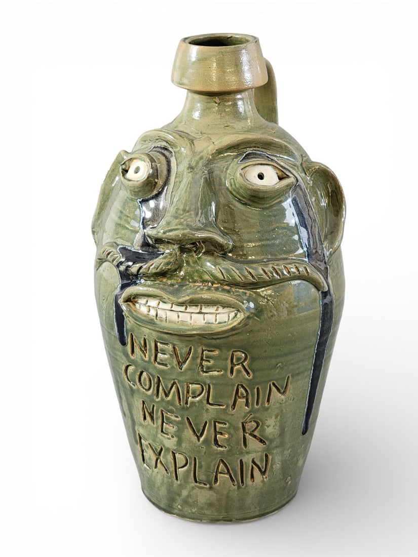 RANDY TOBIAS FACE JUG: Randy Tobias, North Carolina, late 20th-early 21st century, earthenware. A moustached face jug with green glaze. Below face is inscribed "NEVER COMPLAIN NEVER EXPLAIN." Signed on back and bottom. 14"