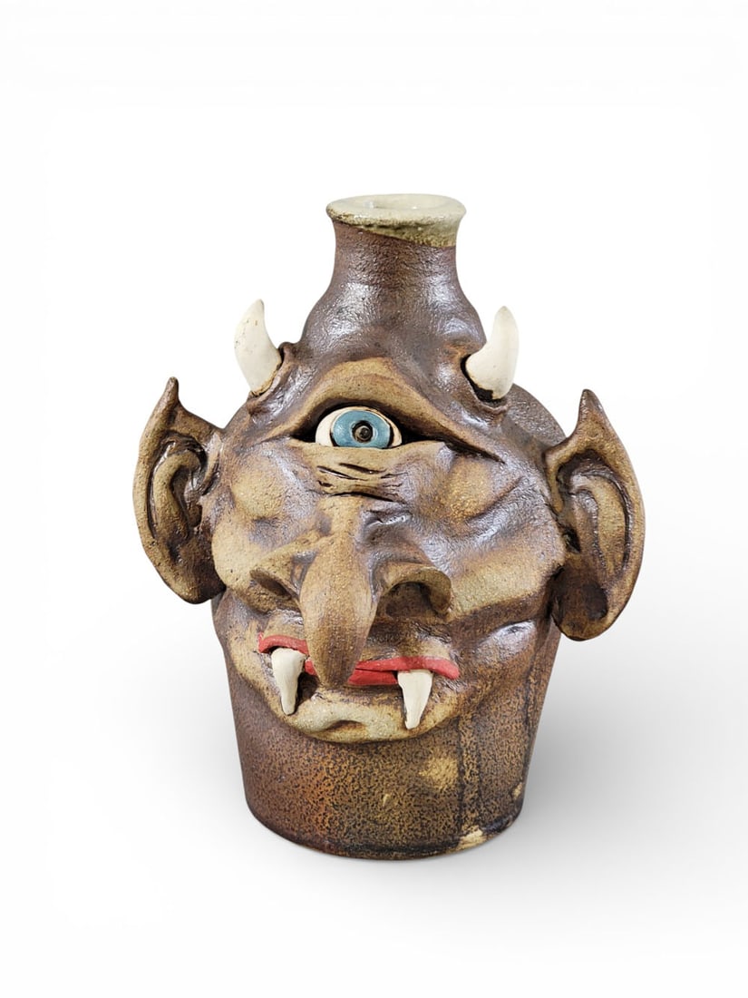 MARK WILLIAMS CYCLOPS FACE JUG: Mark Williams, Georgia, late 20th-early 21st century, earthenware. A cyclops face jug with hooked nose, fangs, horns, and goblin ears. Rear inscribed "IN THE LAND OF THE BLIND THE ONE EYED IS KING." S