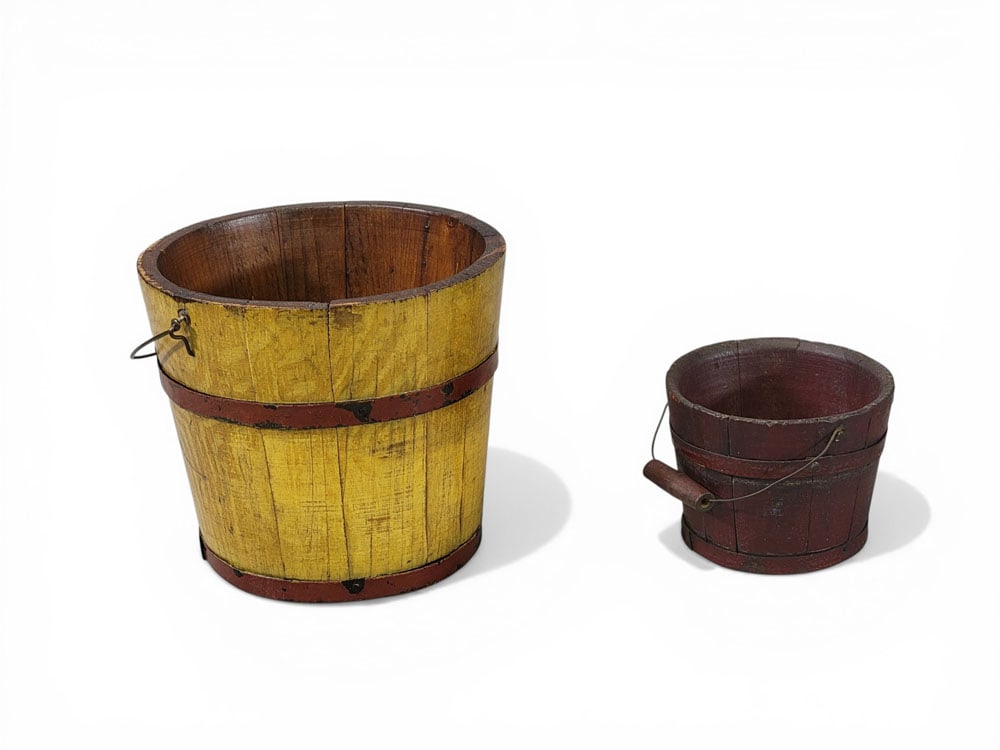 TWO CHILD'S BUCKETS: American, 19th century. Stave-constructed with bail handles. The larger has original yellow paint with red bands, 6.5" high; together with a smaller bucket with original, grungy red paint, 3.5" high.