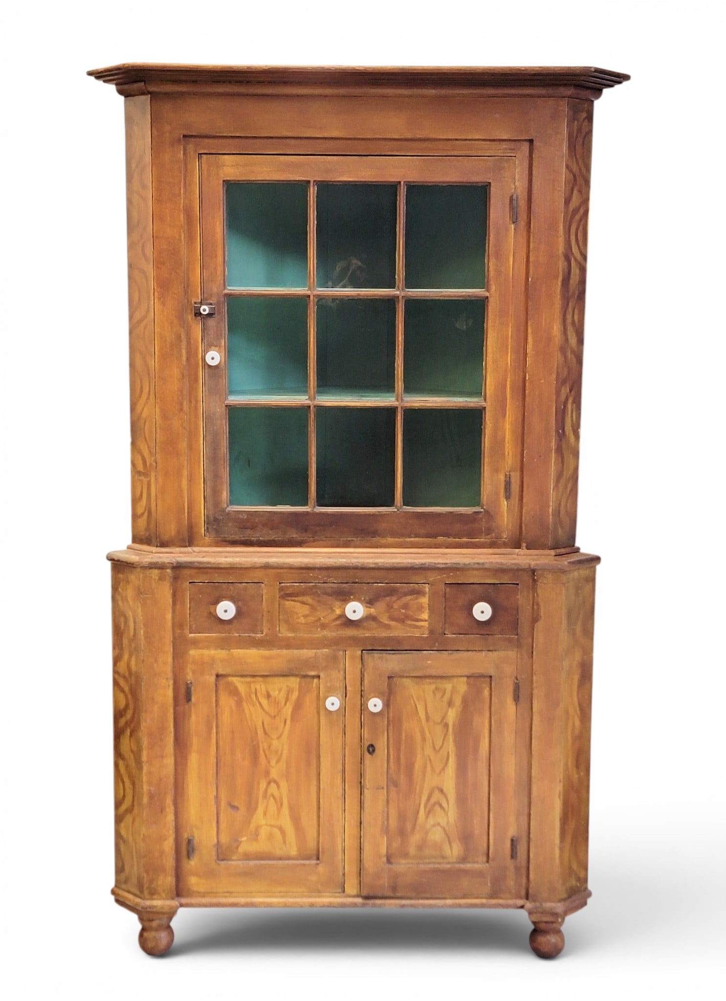 PAINT-DECORATED CORNER CUPBOARD: Pennsylvania, 1840-1860, poplar. Two-piece construction, the upper section with a single, 9-pane door, the lower section with three drawers over two doors, all on turned feet. Retains its original