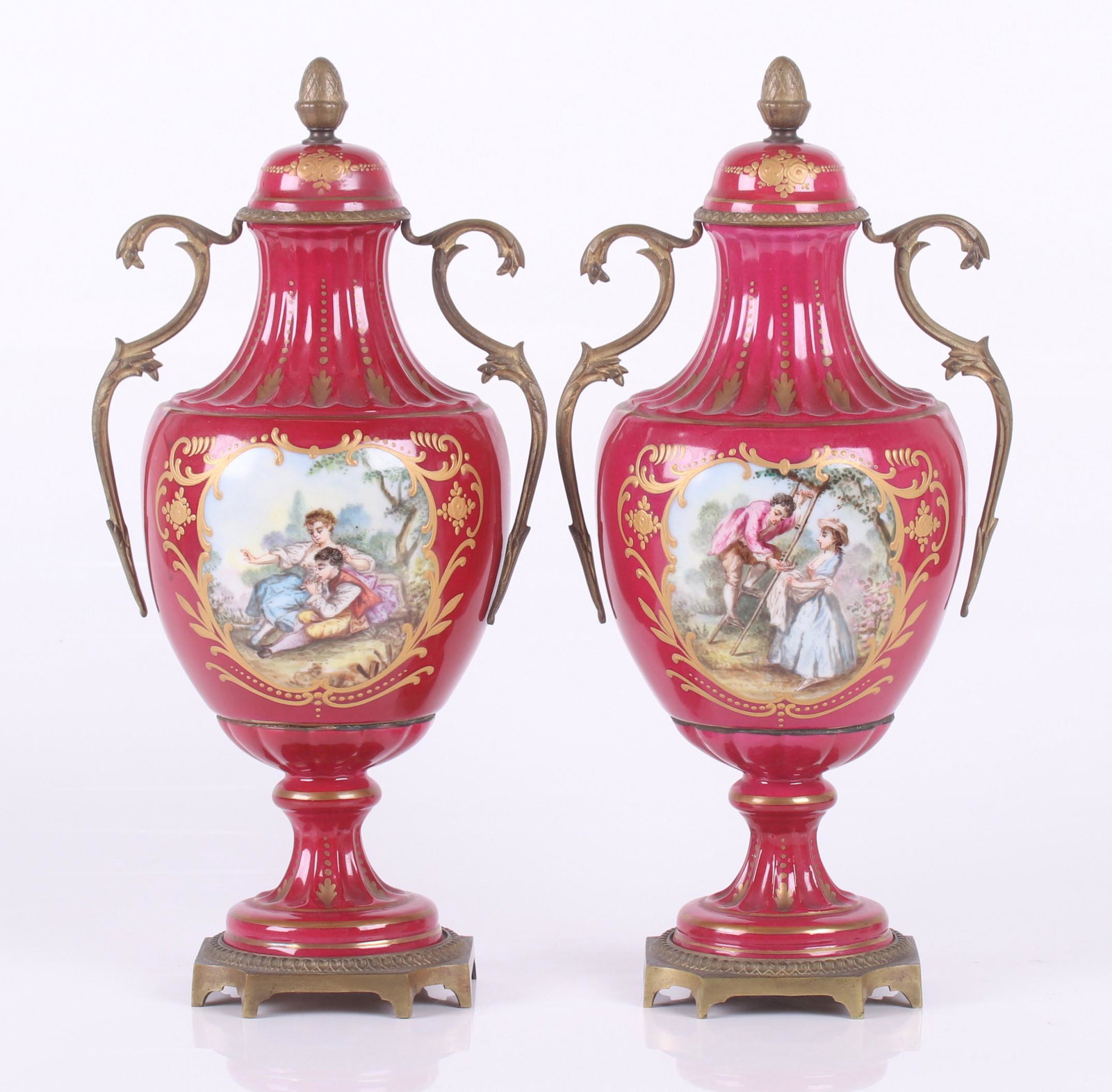 Pair of French Porcelain Urns: A pair of ormolu mounted reeded urns with a crimson ground and gilding, each with hand painted courting scenes, the handles are foliate form and the finials are acorn form; marked as pictured. Dimensi