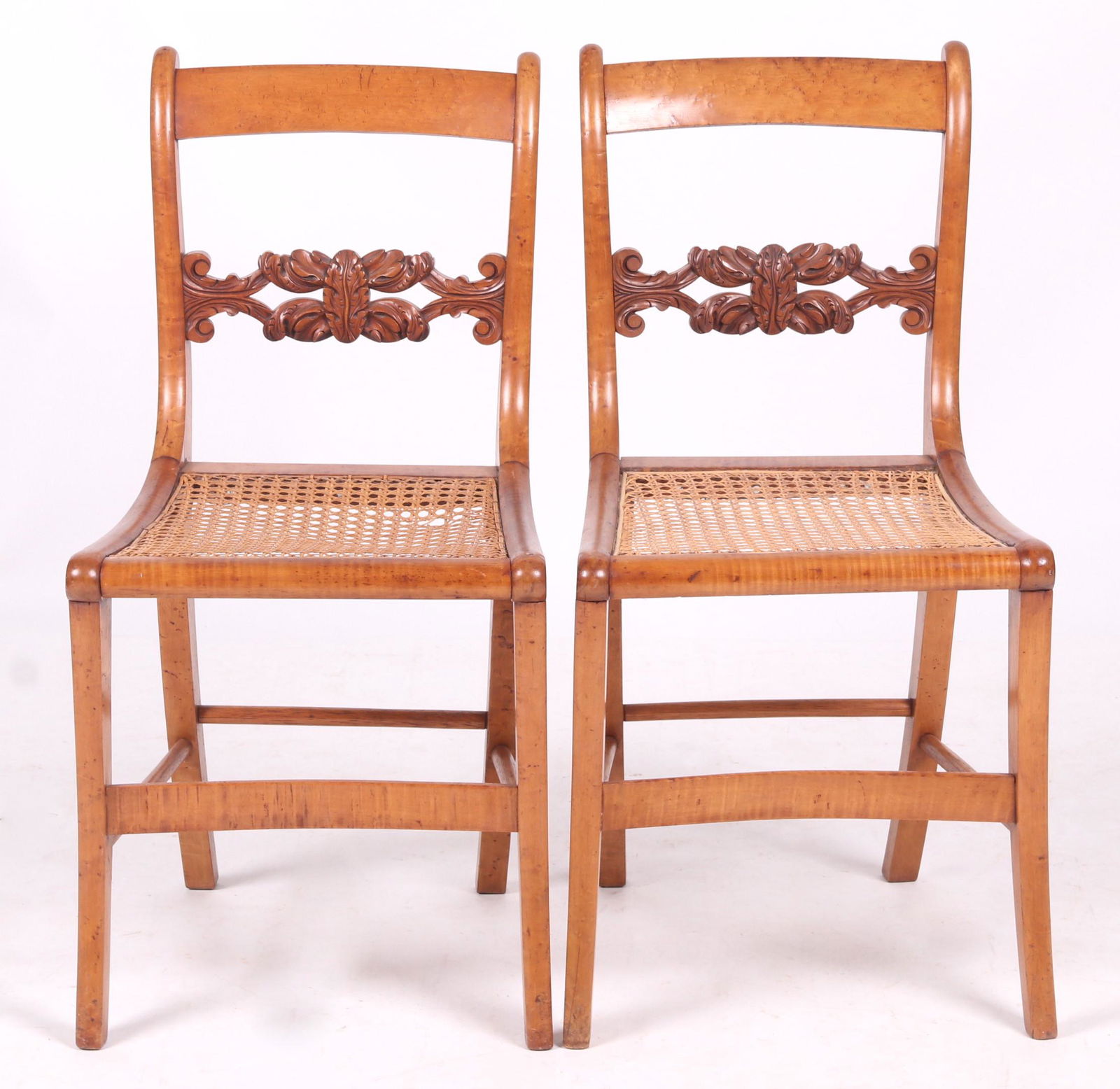A Pair of American Maple Fancy Chairs: Featuring well figured maple, sabre legs, caned seats, and a ladder back with scroll and foliate carved slat. Dimensions:Seat is 16 1/2" x 16". Seat height is 17 1/2". Overall height is 32 1/2". Prove