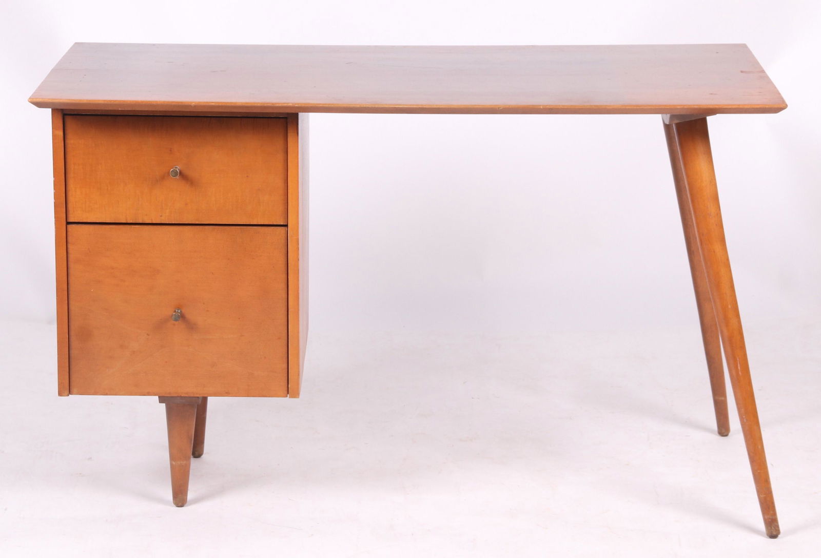 A Mid Century Desk by Paul McCobb: A planner group desk, maple, with long canted legs at one side and short legs at the other below a row of two drawers. All below the rectangular top. Labelled as pictured. Dimensions:24" x 48". Height