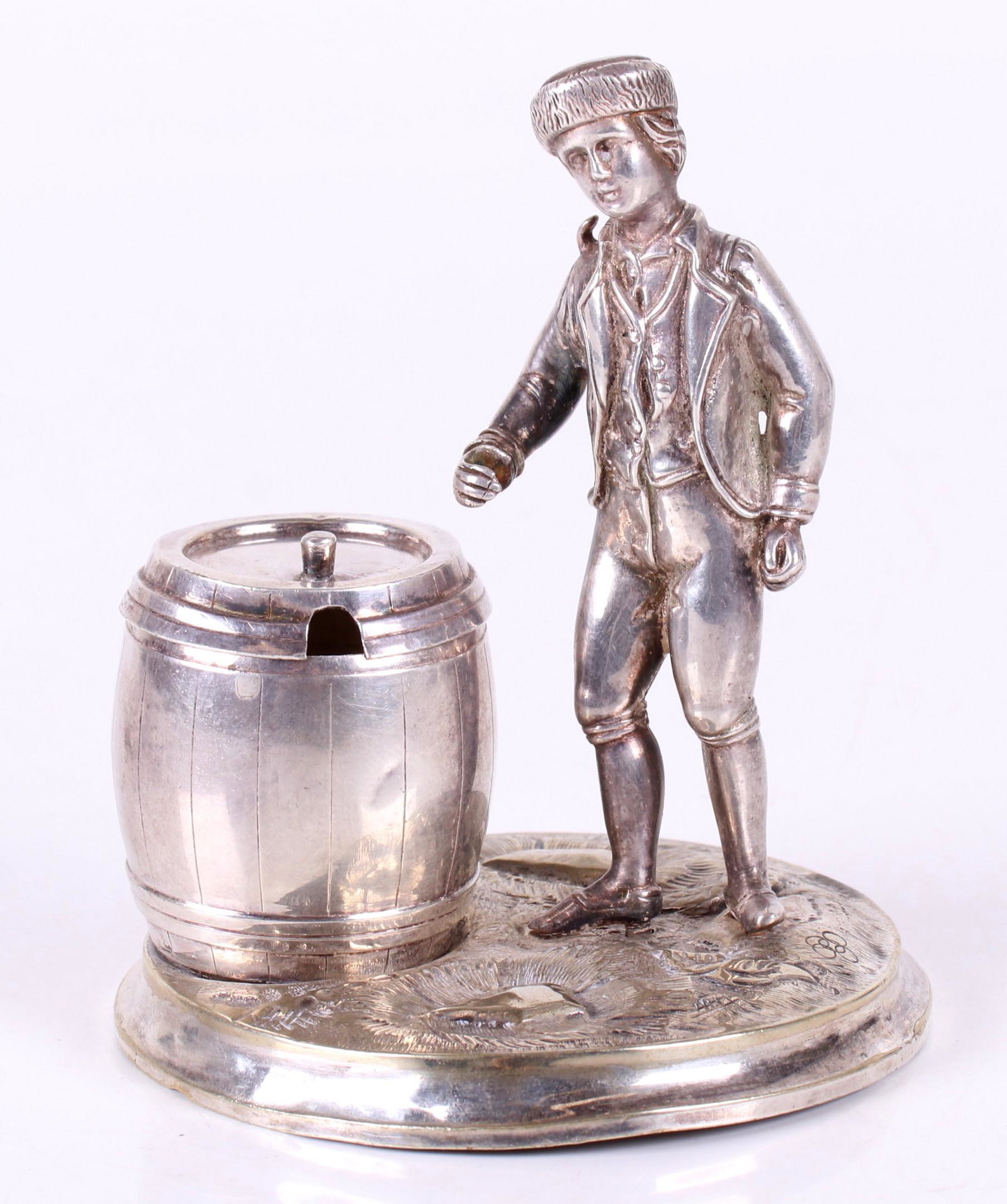 A Continental Silver Figural Mustard Pot: 19th century, depicting a young man wearing a fur cap and standing next to a barrel, both mounted on a grassy mound. Unsigned, the figure and base appear to be silver plated, the barrel, although unma