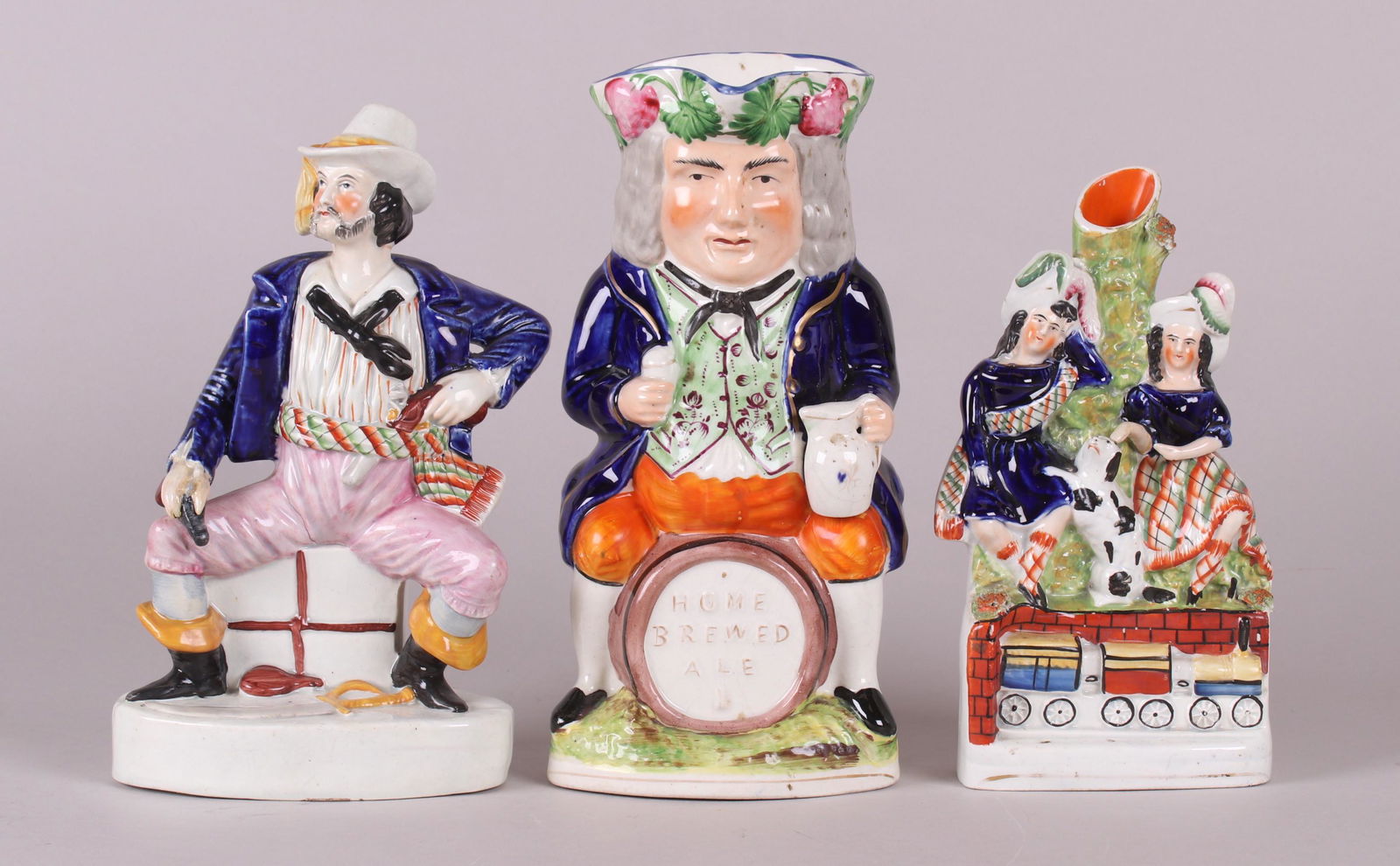 Three Pieces of Staffordshire Pottery: A large Toby pitcher that bears the text "Home Brewed Ale", a figure of a seated man holding pistols and with a sabre at his feet, and a spill vase with two figures, a dog and a toy train set. All mid