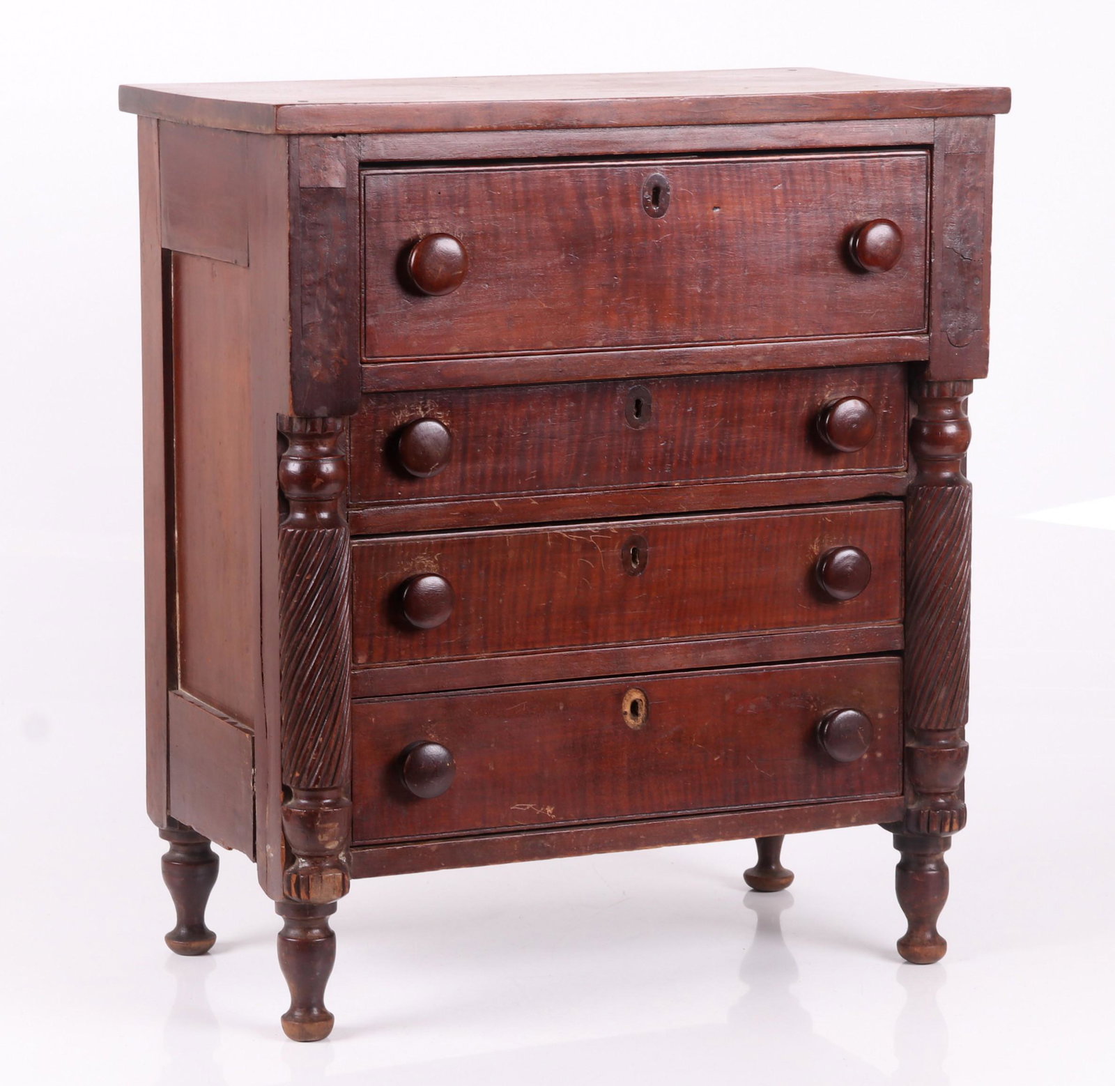 A Child's Size Tiger Maple Chest of Drawers: Circa 1830, with turned feet below a paneled cherry case with four solid tiger maple dovetailed drawers, all below a conforming rectangular top. The piece features half spindle twist-reeded stiles and