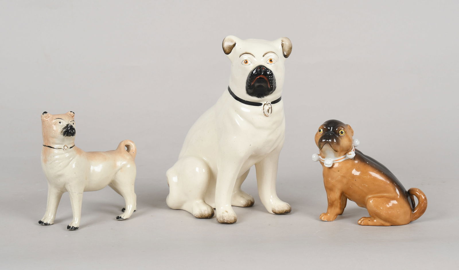 Three English and German Porcelain Pugs, Second Half 19th Century: Three English and German Porcelain Pugs, Second Half 19th Century Comprising: A Staffordshire model of a seated pug and a smaller model of a standing pug with curled tail; and a Dresden-style