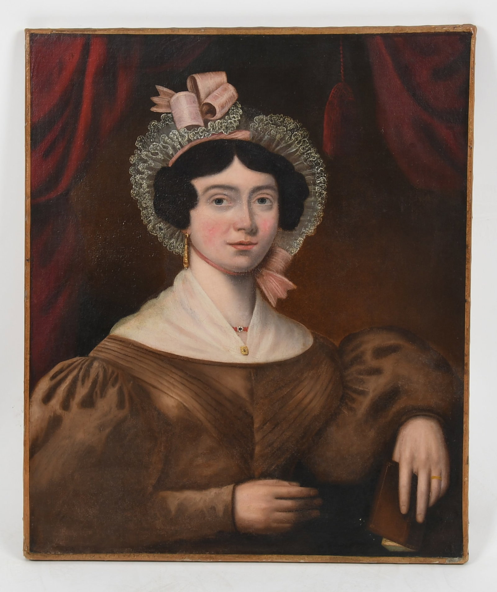 New England School (19th Century), Portrait of a Lady, c. 1830-1840: New England School (19th Century), Portrait of a Lady, c. 1830-1840 Depicted half-length, wearing a lace bonnet with pink ribbon gold earrings and fawn-colored dress holding a book, against a b