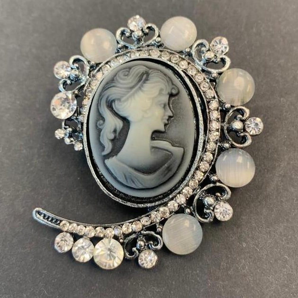 Grey Silhouette Cameo Brooch: Grey Silhouette Cameo Brooch This elegant brooch features a grey cameo depicting the classic silhouette of a lady, beautifully framed by sparkling white crystals and ornate metal carvings. The