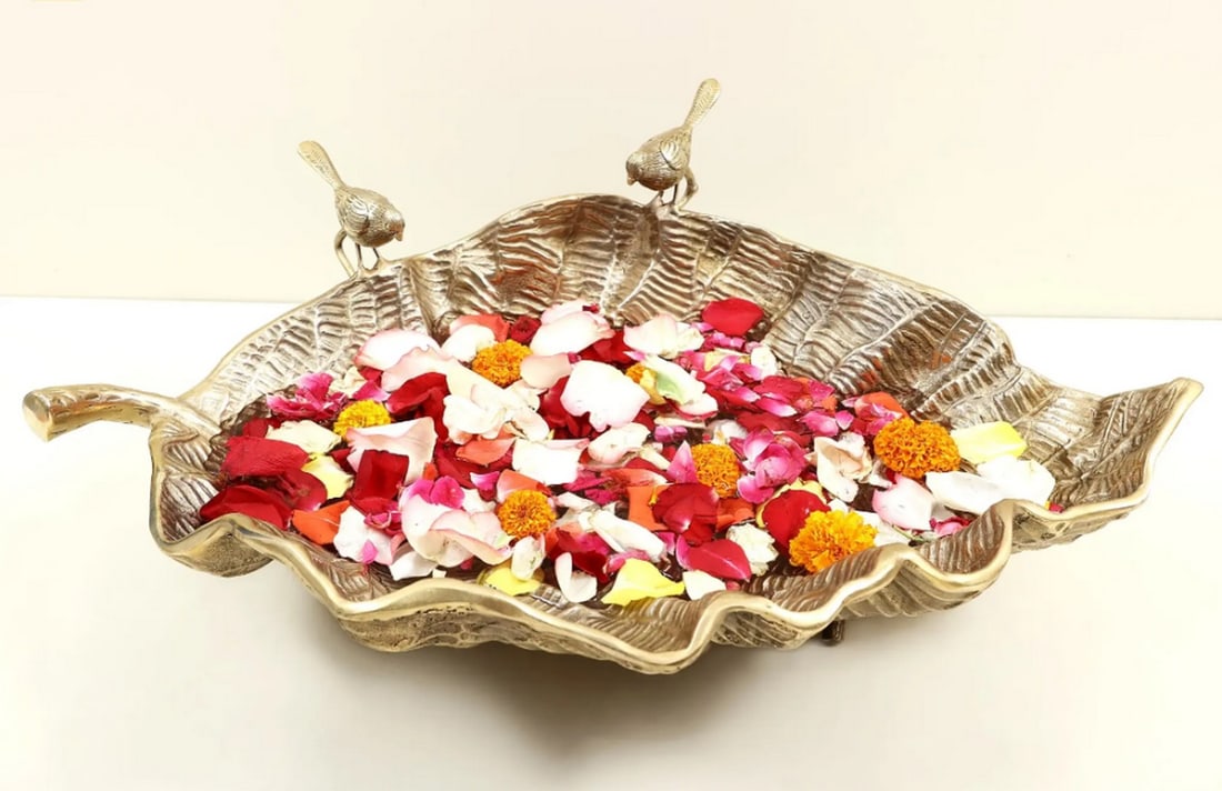 24" Brass Banana Leaf Urli: 24" Brass Banana Leaf Urli Handcrafted from brass, this Urli is shaped in the form of a banana leaf with a textured wavy surface. The design includes two small birds perched on the rim, combining