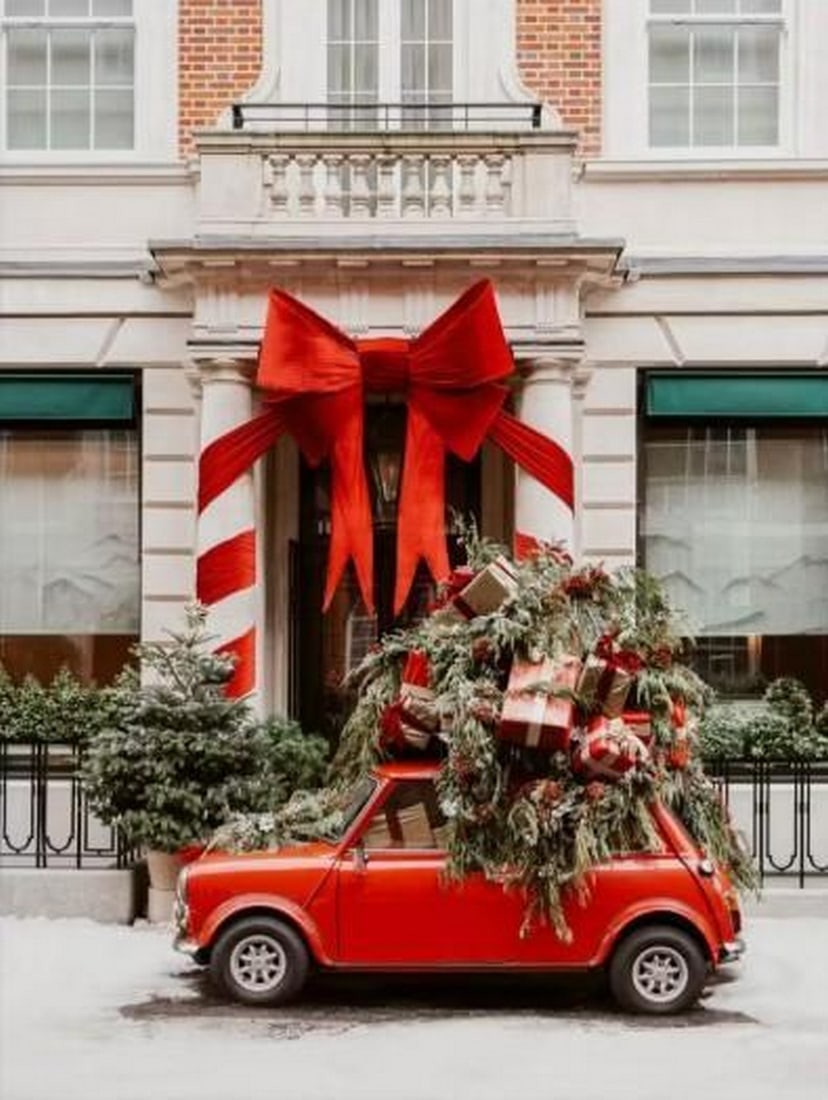 Holiday Stop Canvas Print: Holiday Stop Canvas Print An artwork showing a small red car loaded with gifts and branches, parked beside a brick building. Snow covers the ground, creating a charming and festive Christmas scene wi