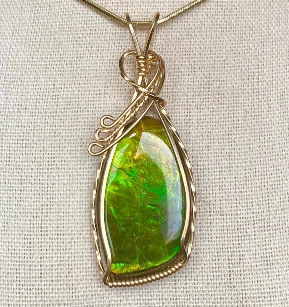 Olive Green Ammolite Pendant in Gold-Filled Wire: Olive Green Ammolite Pendant in Gold-Filled Wire This ammolite gemstone, measuring 27mm x 16mm, features a shiny resin coating and is wrapped in 14K gold-filled wire. The pendant comes on an 18" g