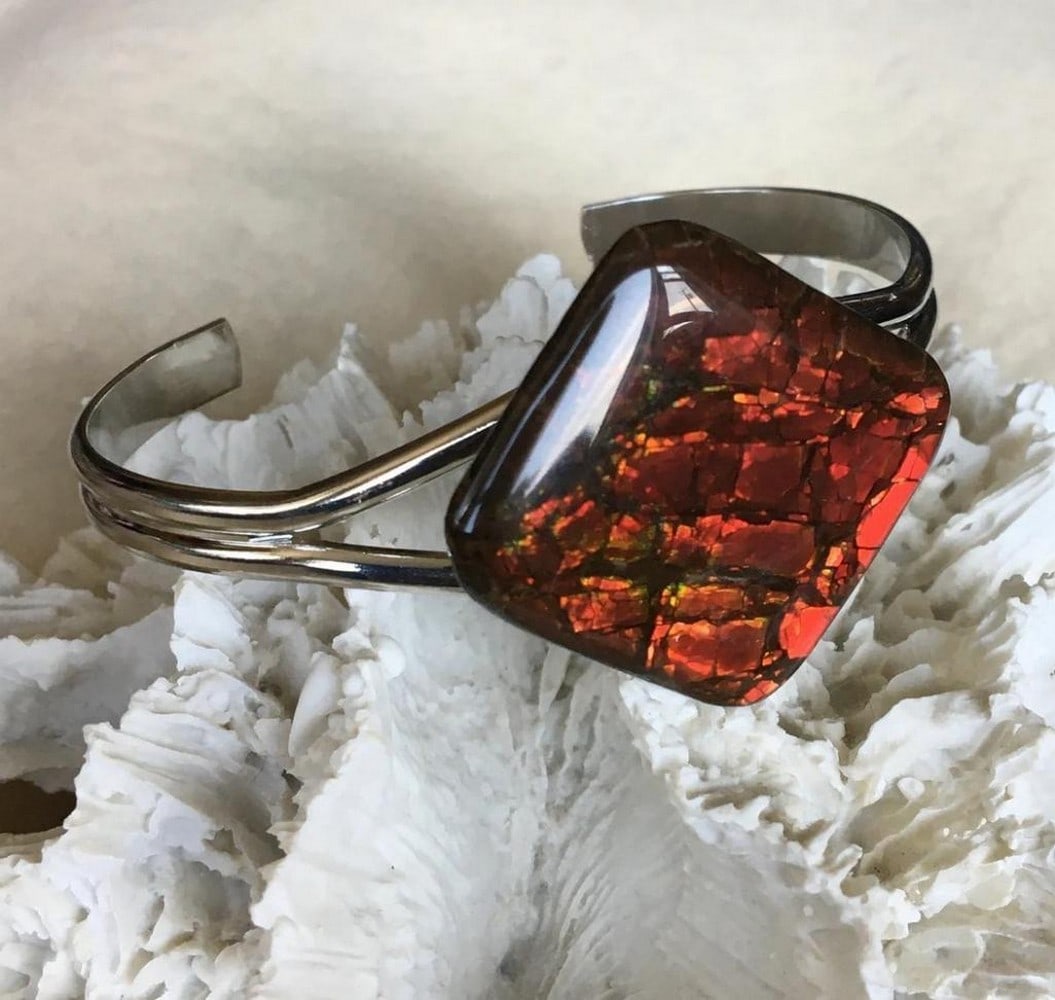 Crimson Aurora Ammolite Cuff: Crimson Aurora Ammolite Cuff This cuff bracelet features a red ammolite gemstone in a stained glass pattern, with a freeform shape measuring 35mm x 34mm. The resin-coated gemstone displays hints of