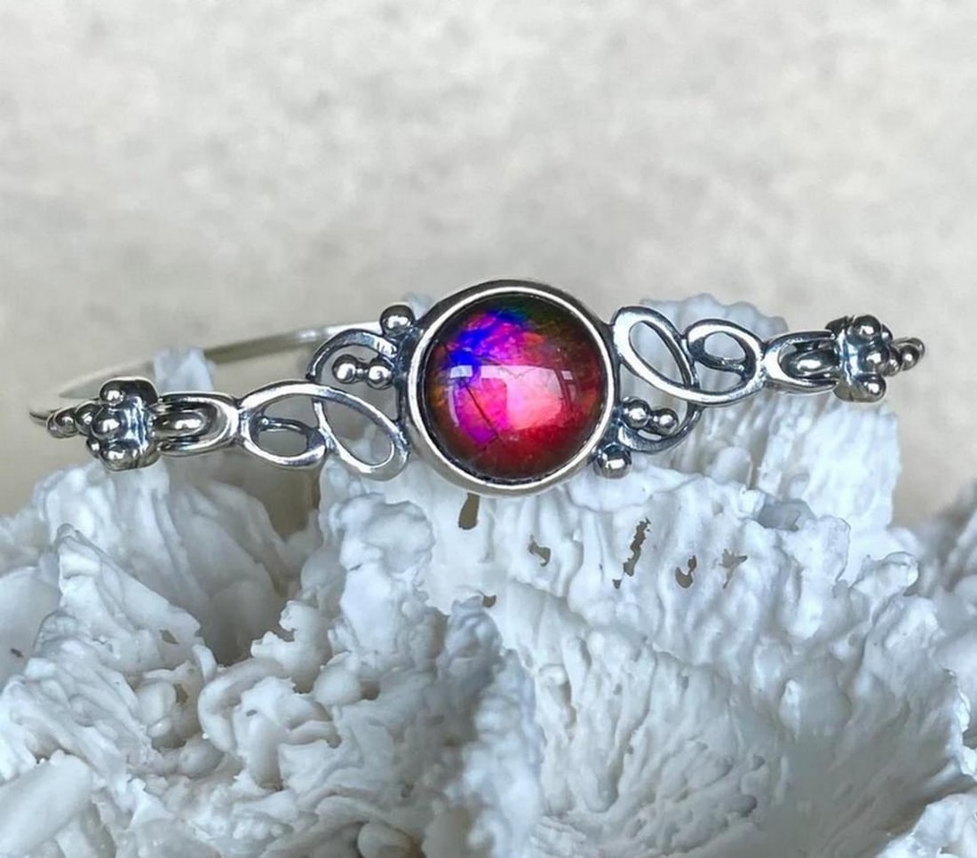 Mystic Shift Ammolite Cuff Bracelet: Mystic Shift Ammolite Cuff Bracelet This elegant bangle features an ammolite triplet with a quartz cap. The gemstone primarily displays a rich red hue, with subtle hints of pink and purple visible fro