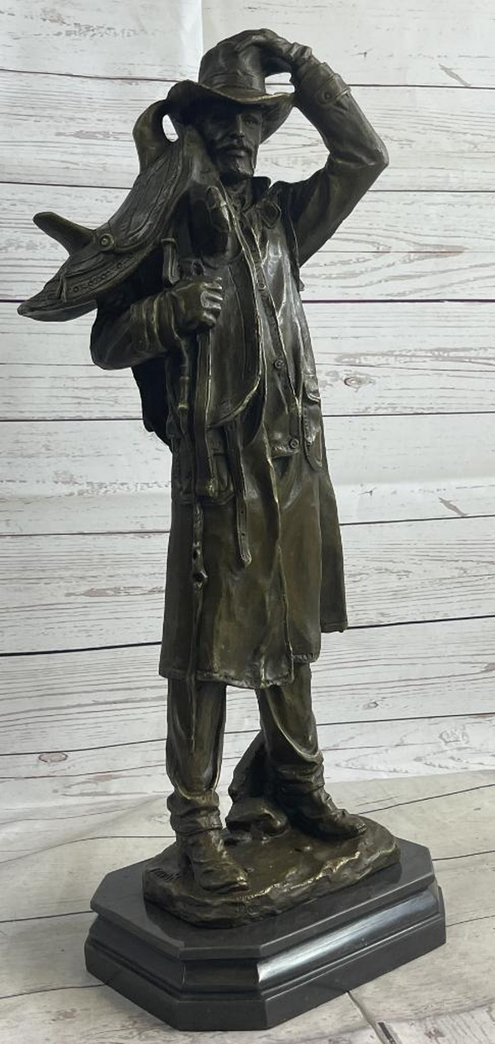 Country Western Cowboy Hauling Saddle Bronze Sculpture Statue Art Decor: This is a sculpture of a classic cowboy. He wears his cowboy hat and his trench coat. On his feet her wears boots which his jeans cover in between his legs is a bunch of twigs that were gathered for f