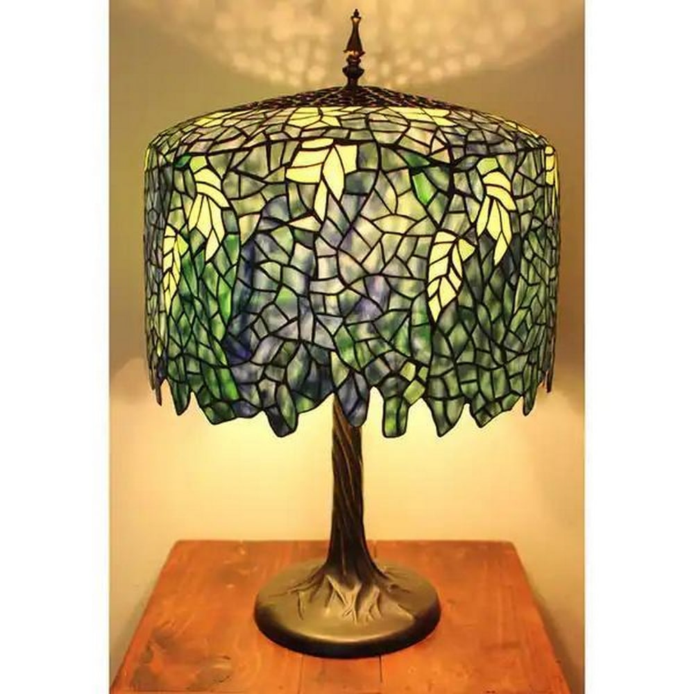 Blue Wisteria Tiffany Style Table Lamp with Sculpted Tree Trunk Base: Blue Wisteria Tiffany Style Table Lamp with Sculpted Tree Trunk Base Bring timeless beauty and warm ambiance to your space with this Blue Wisteria Tiffany Style Table Lamp. The stained glass shade is 
