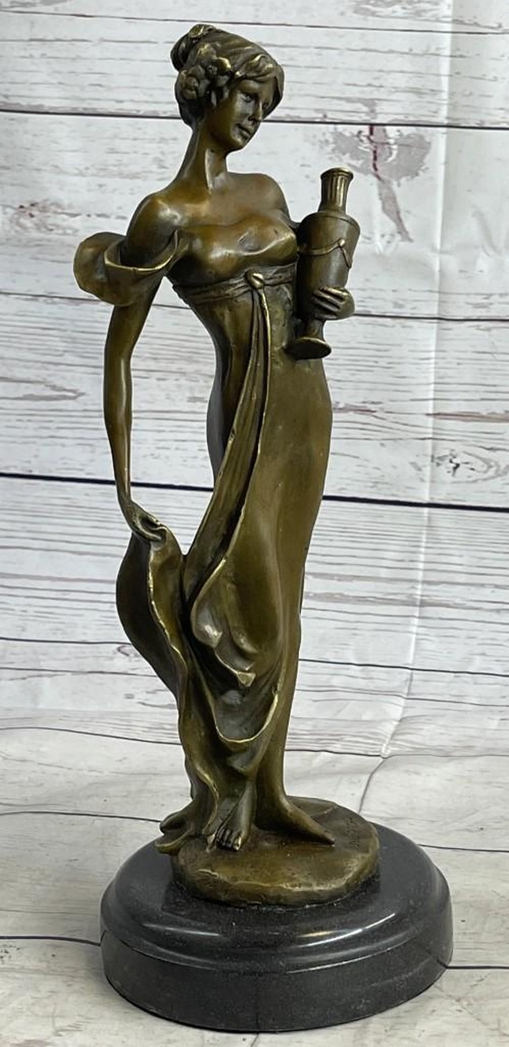 Woman Holding Vase Bronze Sculpture: Woman Holding Vase Bronze Sculpture This classical maiden stands in an upright position, cradling an empty vase at her side with one hand, holding the bottom of her dress of the ground with her other.