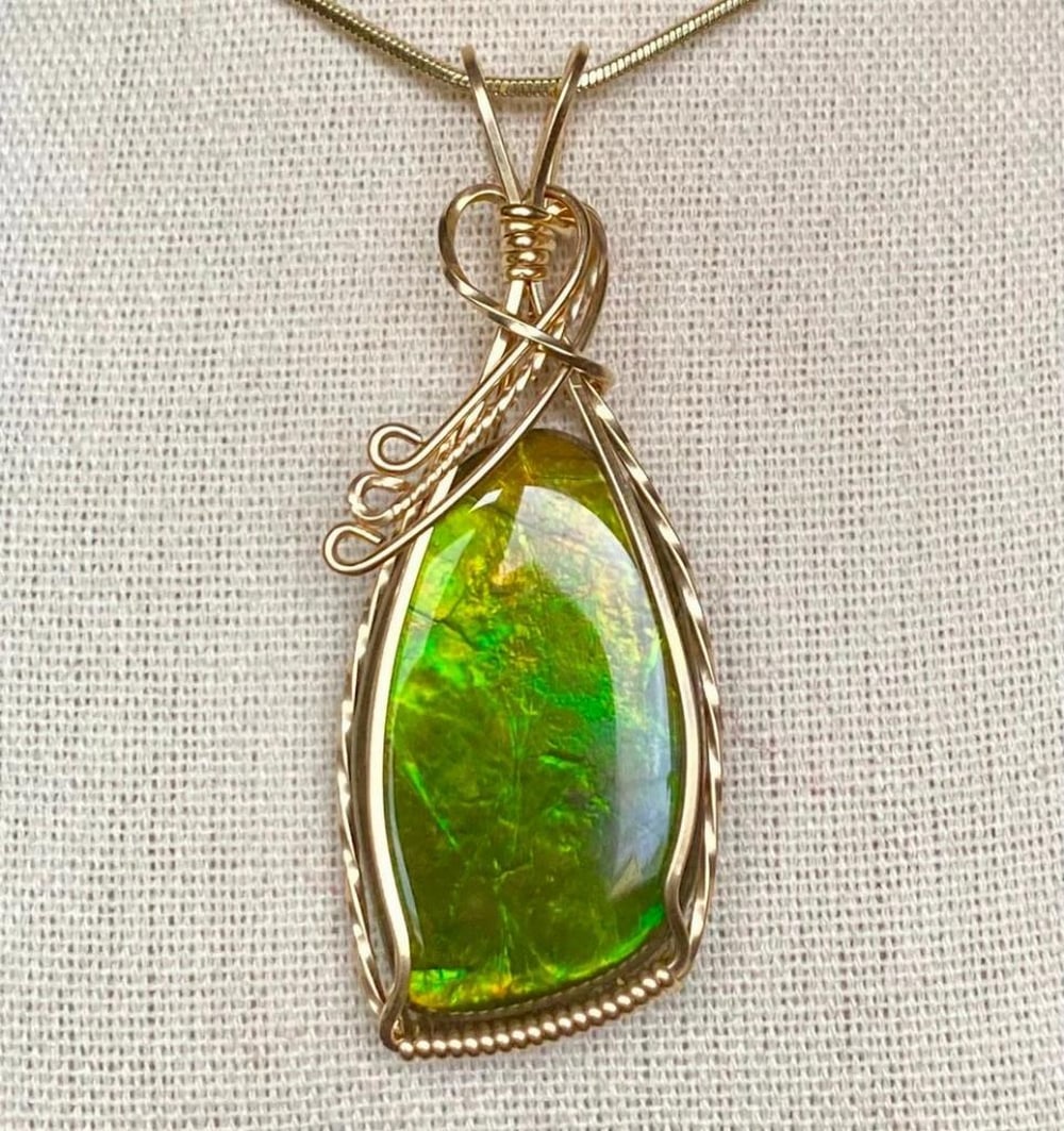 Olive Green Ammolite Pendant in Gold-Filled Wire: Olive Green Ammolite Pendant in Gold-Filled Wire This ammolite gemstone, measuring 27mm x 16mm, features a shiny resin coating and is wrapped in 14K gold-filled wire. The pendant comes on an 18"
