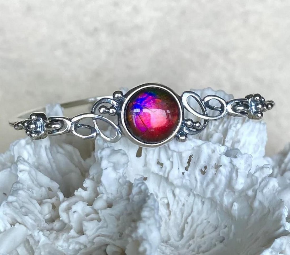 Mystic Shift Ammolite Cuff Bracelet: Mystic Shift Ammolite Cuff Bracelet This elegant bangle features an ammolite triplet with a quartz cap. The gemstone primarily displays a rich red hue, with subtle hints of pink and purple visible