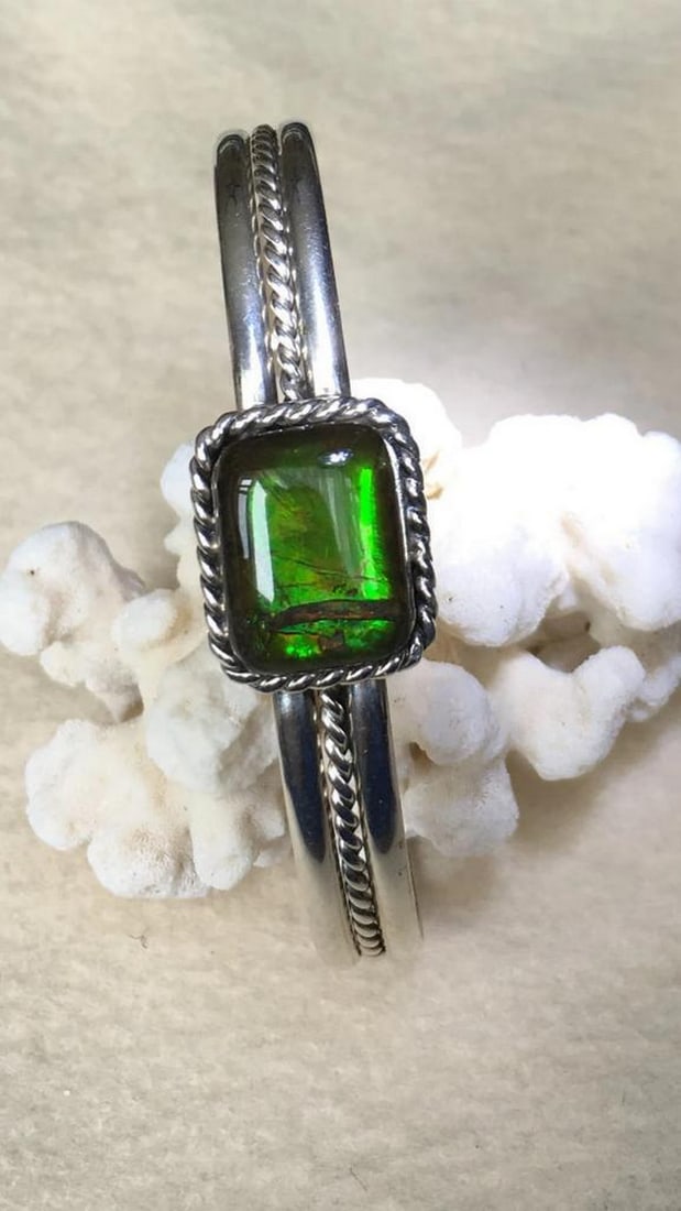 Emerald Aurora Ammolite Cuff: Emerald Aurora Ammolite Cuff This sterling silver cuff bracelet features a green ammolite gemstone set on a rope-edged plate, showcasing its natural beauty. Upon close inspection, a small section of t