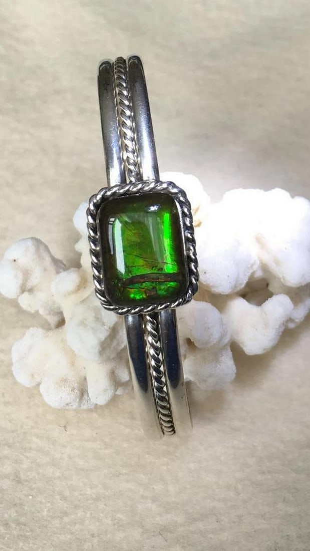 Emerald Aurora Ammolite Cuff: Emerald Aurora Ammolite Cuff This sterling silver cuff bracelet features a green ammolite gemstone set on a rope-edged plate, showcasing its natural beauty. Upon close inspection, a small section of