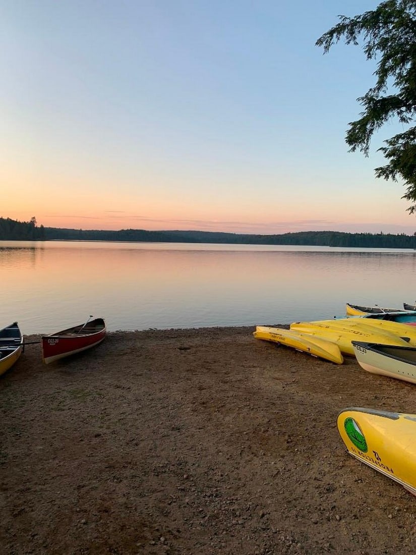 Algonquin Evening Reflections: Algonquin Evening Reflections A tranquil photograph taken from the sandy shore of a lake in Algonquin Provincial Park, where canoes rest quietly by the water's edge as the sun sets beyond the distant