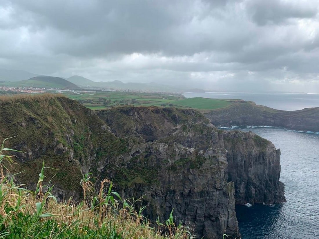 Misty Coastal Cliffs: Misty Coastal Cliffs The photograph showcases a rocky cliffside from the vantage point of the edge, with glimpses of the ocean below and distant hills and a quaint village shrouded in fog on the horiz