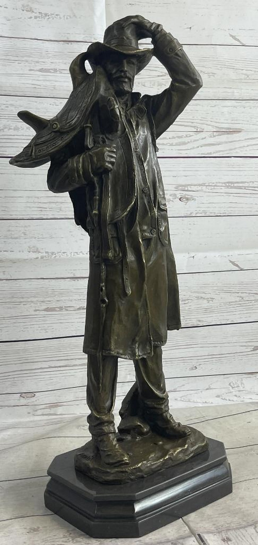 Country Western Cowboy Hauling Saddle Bronze Sculpture Statue Art Decor: This is a sculpture of a classic cowboy. He wears his cowboy hat and his trench coat. On his feet her wears boots which his jeans cover in between his legs is a bunch of twigs that were gathered for f