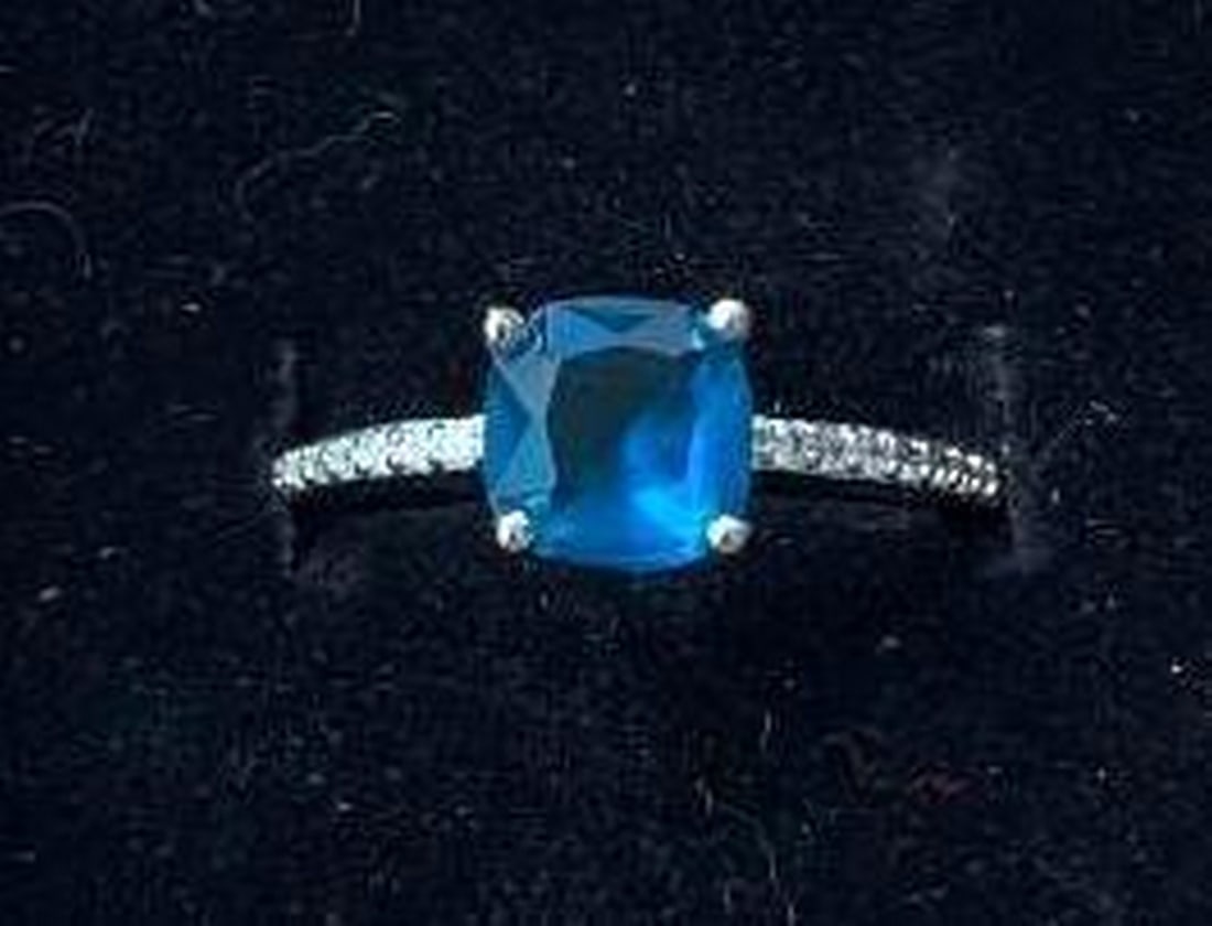 Sterling Silver Ring with Cushion-Cut Blue Stone: Sterling Silver Ring with Cushion-Cut Blue Stone This striking sterling silver ring showcases a captivating cushion-cut blue stone at its center, radiating a rich medium blue hue. Flanking the blue st