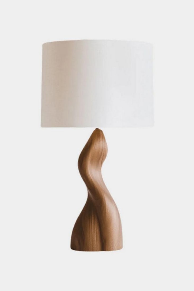 Contemporary Flowing Wood Table Lamp: Contemporary Flowing Wood Table Lamp This table lamp features a solid wood base carved into a flowing, sculptural form with smooth curves and visible natural grain. The organic silhouette is paired
