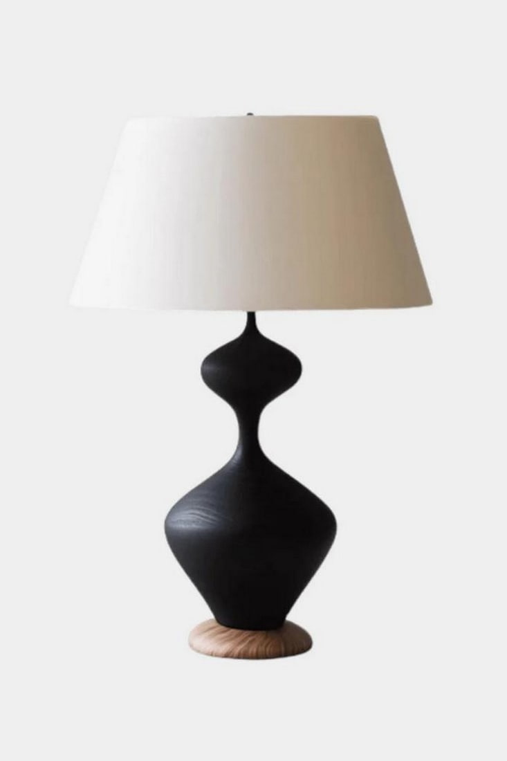 Hourglass Silhouette Table Lamp: Hourglass Silhouette Table Lamp Sleek and sculptural, this handcrafted table lamp features a curvaceous black base with an hourglass-inspired form, resting on a circular wooden platform. The wide