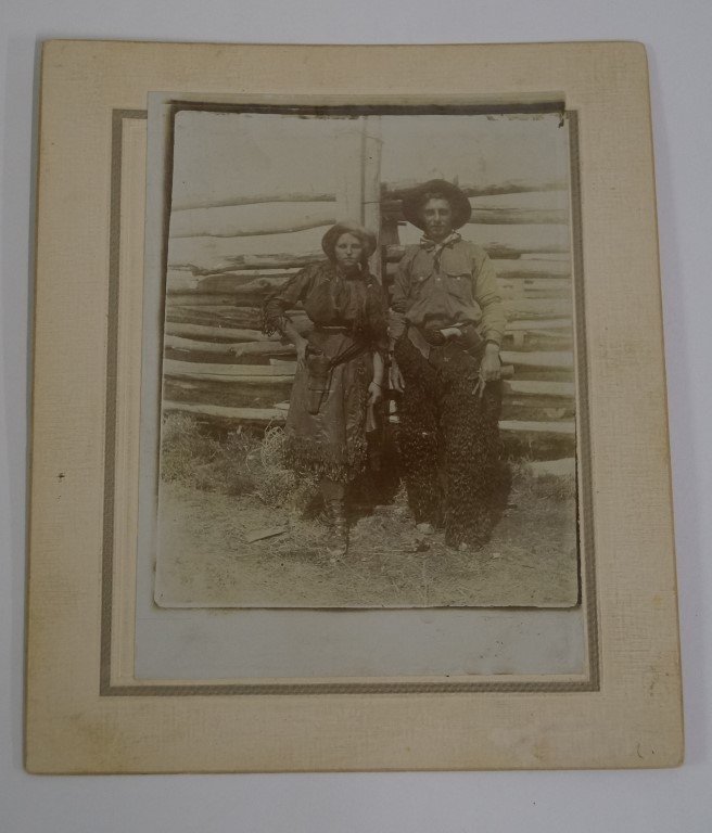 "Annie Oakley" Cowboy and Cowgirl Photograph: "Annie Oakley" type cowgirl in bonnet with pistol in holster poses with a cowboy in "fur" chaps with his ivory handled gun in holster. 4x6" on larger cream colored cardboard.