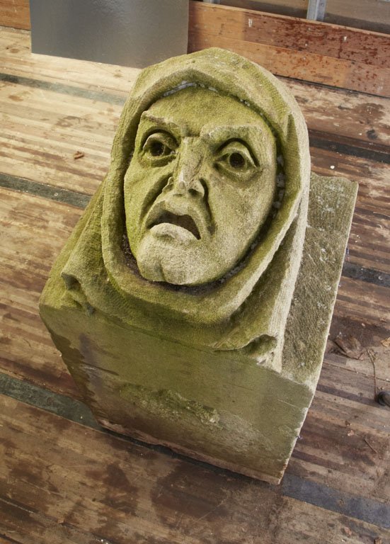 CARVED FIGURAL HEAD ON LIMESTONE, C. 1900: Perhaps portraying a monk or a clergyman, this limestone head peers out from a hooded cloak. The eyebrows are exaggerated to depict shock or surprise and the mouth agape. The eyes are heavily-lidded a