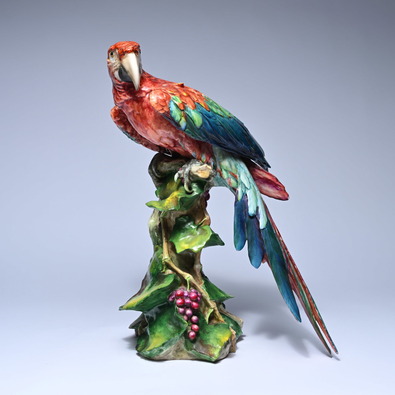 Polychrome painted parrot (macaw) in glazed ceramic on a branch among lush green leaves and a bunch (1 of 9)