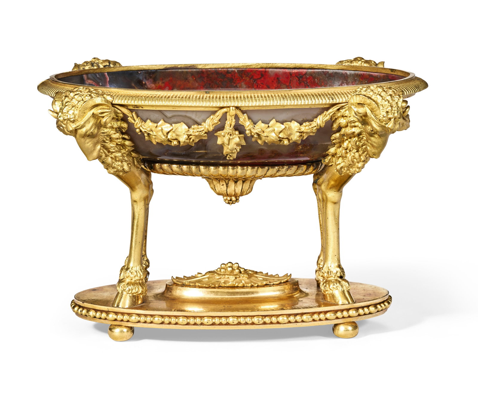 A LATE LOUIS XVI ORMOLU-MOUNTED AGATE TAZZA CIRCA 1785: Details In matte and burnished gilding, the oval bowl of vermilion-veined grey agate, with a reeded border supported by ram monopodia joined by ivy festoons, on an oval base centered by a patera 73