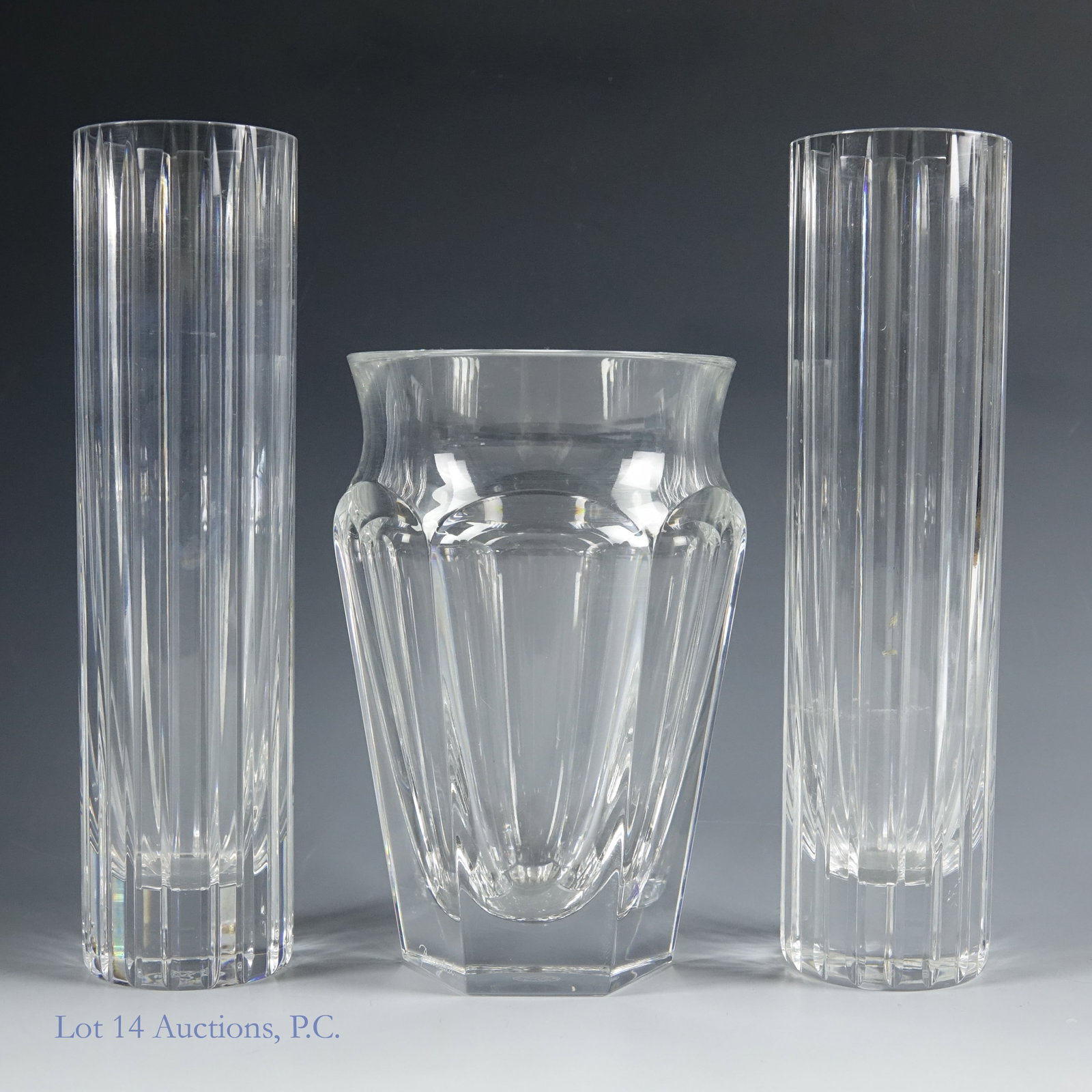 Three Baccarat Crystal Vases: Three crystal glass vases from Baccarat. The first is an approximately 5" tall vase in the Nelly style. The other two are a matching set of cylinder Harmonie vases, each one approximately 6.75" tall. 