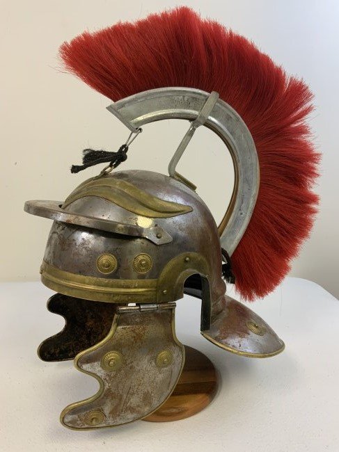 ROMAN CENTURION STEEL HELMET REPLICA WITH RED PLUMAGE: A steel replica of the military head armor worn by Roman centurions dating back to the reign of Julius Caesar, this weathered helmet features brass accents, moveable cheek plates, and a decorative red