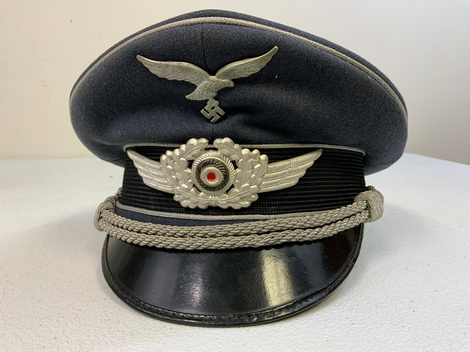 WWII GERMAN LUFTWAFFE OFFICER VISOR CAP: WWII GERMAN LUFTWAFFE OFFICER VISOR CAP . BLUE WOOL BODY WITH ALUMINUM INSIGNIA INSTEAD OF EMBROIDERED. POSSIBLE OFFICER SCHOOL CANDIDATE . GOOD WORN CONDITION . MAKER MARK ON THE SWEAT DIAMOND.