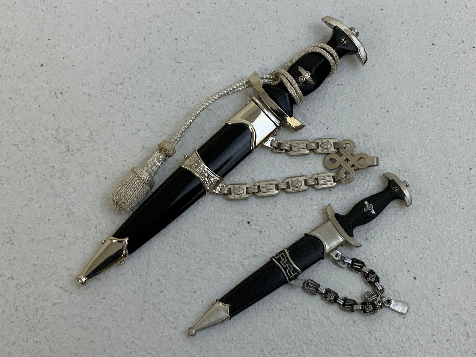 SS OFFICER MINIATURE DAGGERS LOT OF 2 CHAINED: SS OFFICER MINIATURE DAGGERS LOT OF 2 CHAINED. 7" LONG WITH 4 1/2" LONG BLADE AND 4 1/2" WITH 3" LONG BLADE. SMALLER ONE HAS A CHAIN LING MISSING.