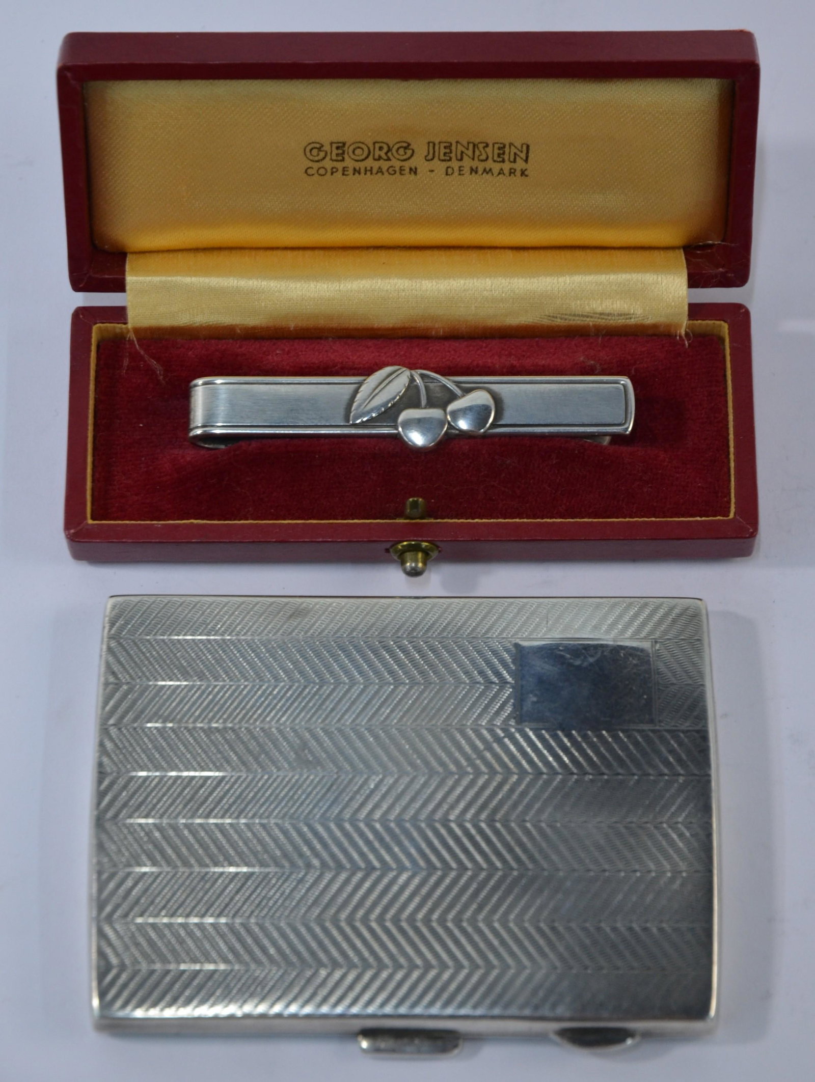 A Georg Jensen tie-clip and English Art Deco cigarette: Georg Jensen:Â a cased Sterling tie-clip mounted with cherries, inscribed on reverse 'Cherry Heering' (mark used 1945 to present), to/w an Art Deco engine-turned herringbone patterned cigarette cas